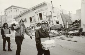 Three CCC Corpsmembers carry supplies in front of leveled building on San Francisco street 1989