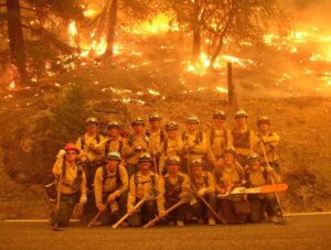 Fire crew Corpsmembers pose in equipment while hillside behind them is engulfed in flames