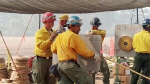 Corpsmembers in fire gear use hose rolling machine to roll hose at fire camp