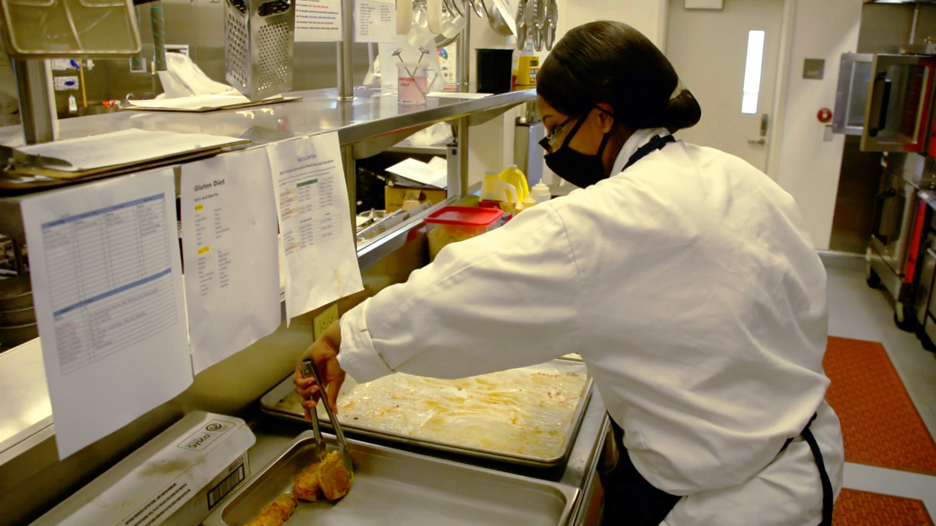 Female in kitchen using tongs to pick up food to move to a tray