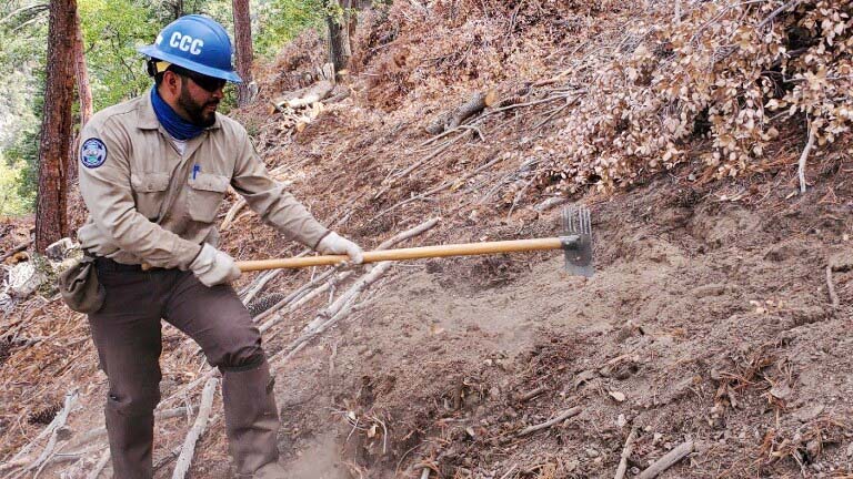 male corpmember in ppe uses mcleod to rake ground debris around burn pile