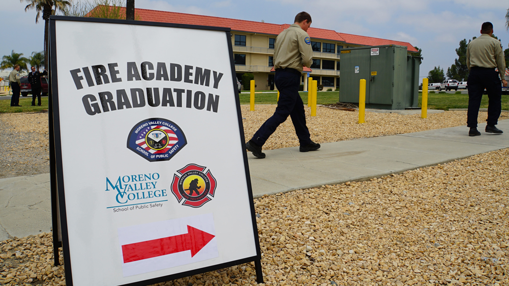 sign reading fire academy graduation with an arrow in foreground as corpsmembers walk through background