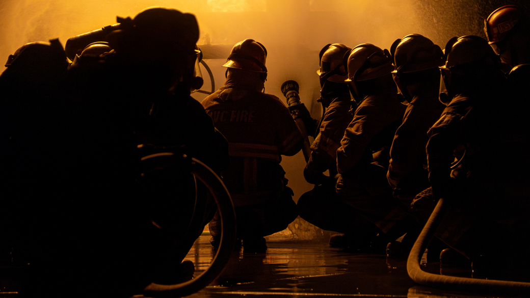 firefighters illuminated by flame as they crouch down with fire hose spraying water