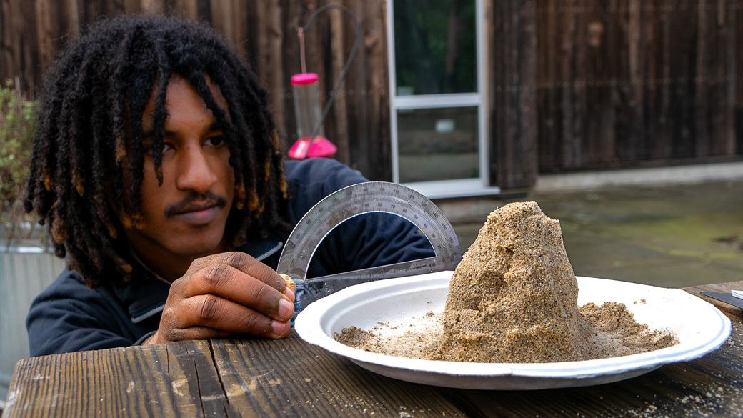 male holding plastic protractor at bench table looking at a pile of sand