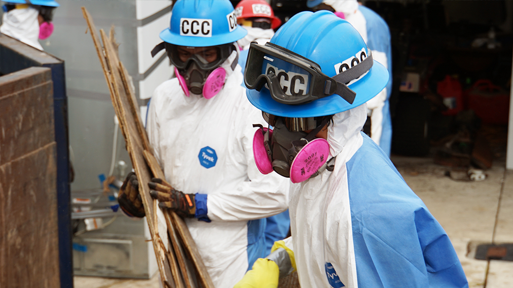 corpsmembers in helmets, respirators and tyvek haul debris to a trash bin