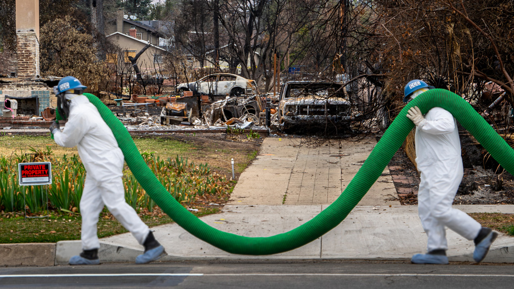 corpsmembers in full PPE haul silt sock in front of a burned home in Altadena