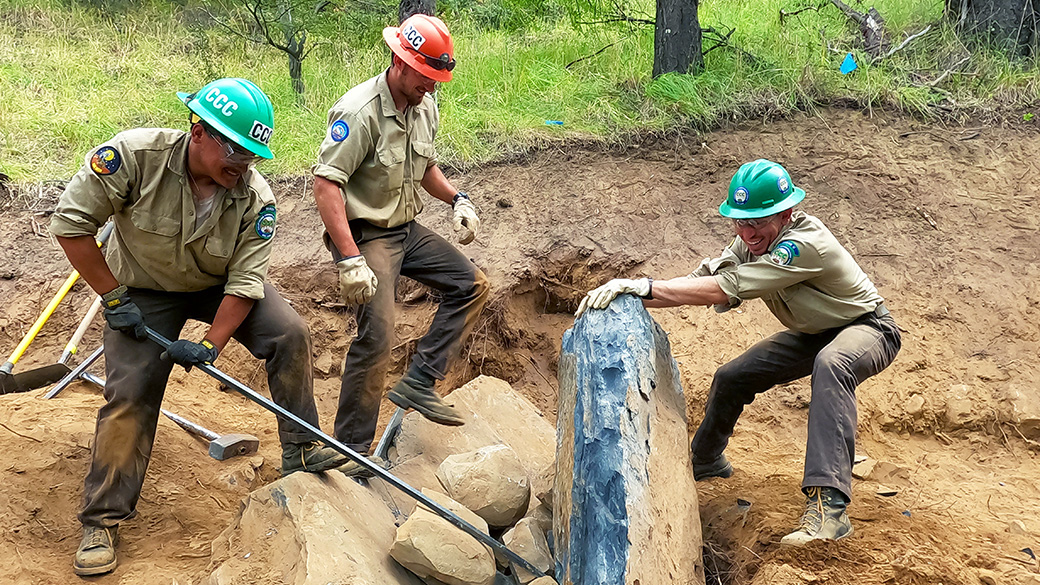 three corpsmembers working together to remove a giant rock in a trail by using rock bars and their body weight