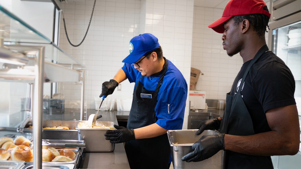 corpsmember stirs in a serving tray with another corpsmember arriving next to him with another serving tray