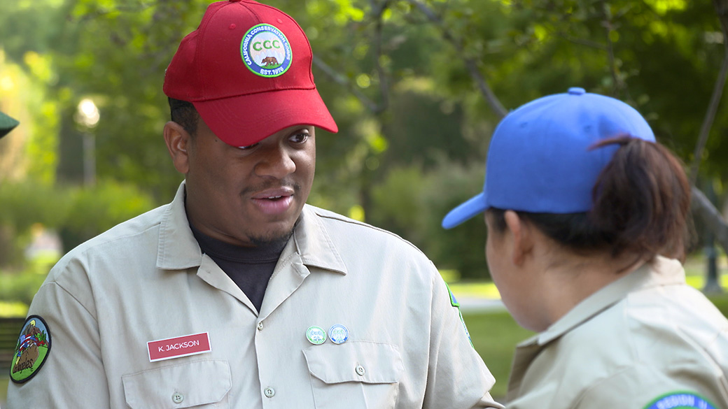 male corpsmember in red hat talking to female corpsmember in blue hat in a park